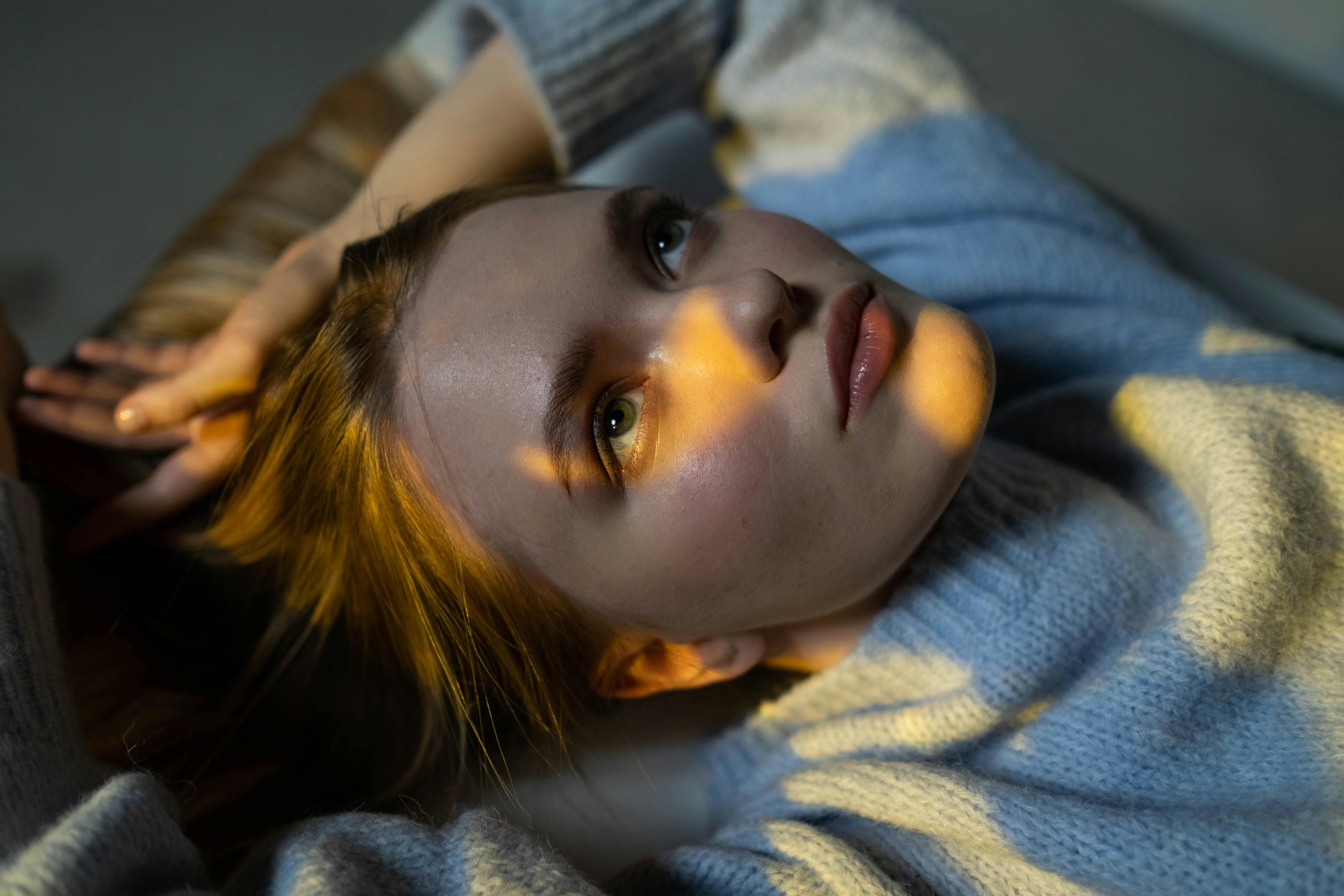 Woman lying down in soft light, gazing upward thoughtfully, with a shadow cast across her face and wearing a cozy blue sweater.