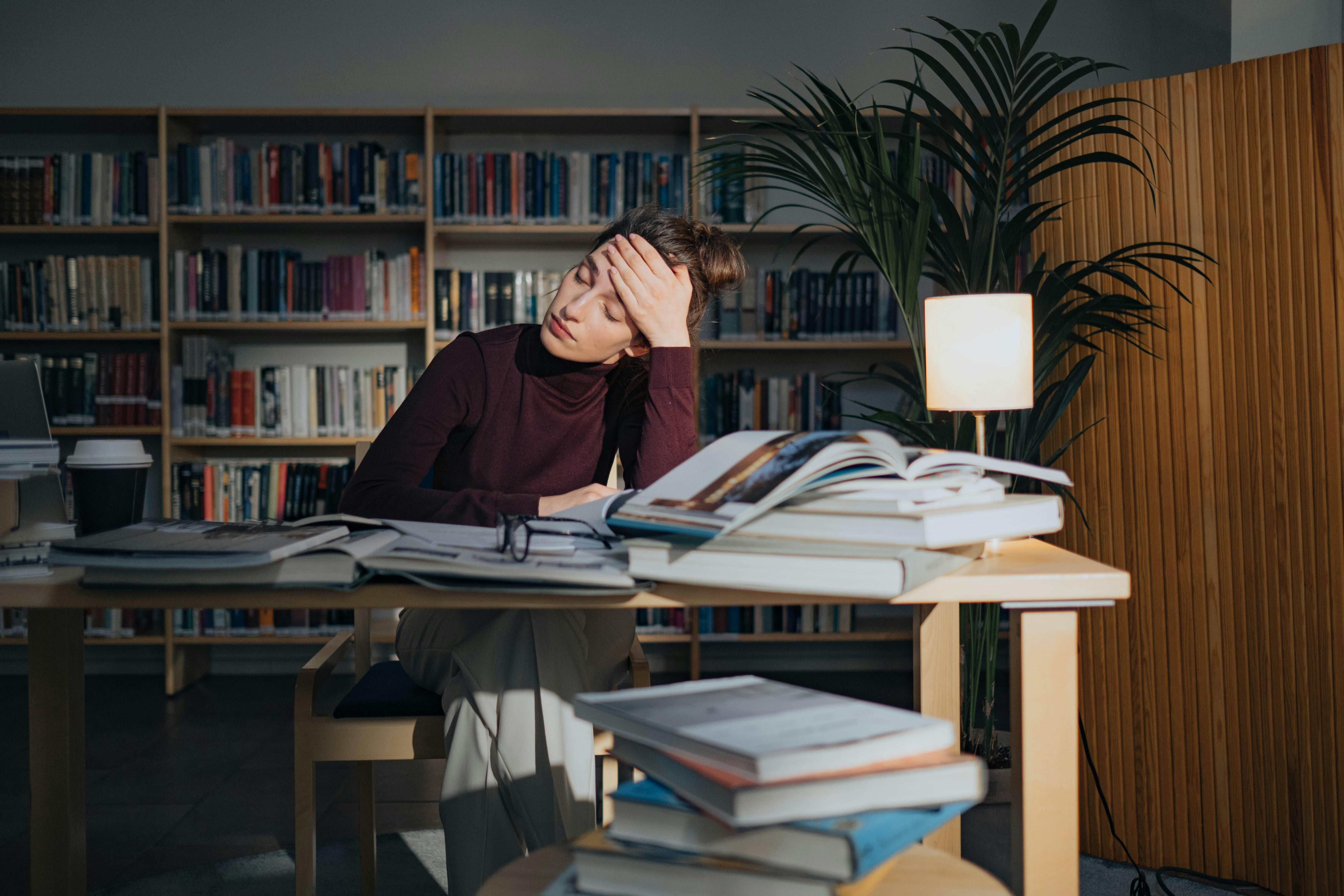 A tired woman sits at a desk in a library, surrounded by open books and papers, resting her head on her hand.